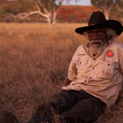 A man reclines on the ground, wearing a dark hat in the outback