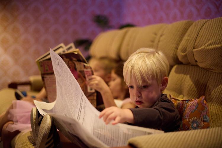 A young child with blonde hair and a brown jumper sits on a 1970s style couch and holds a newspaper in their hands and looks at it with interest