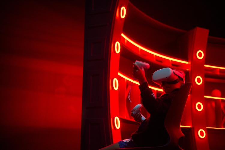 A young person sits on a chair and wears a white VR headset while holding two white VR controllers. They are backlit by the dramatic red lighting emanating from a sci-fi-styled bar area.