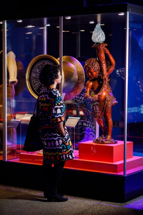A person with curly black hair and a colourful geometric patterned dress stares intently at a statue of a cherub holding a flame in a Museum showcase.