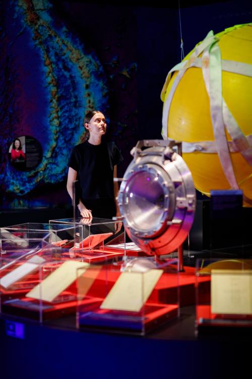 A person in a black t-shirt with their brown hair in a ponytail stares intently at a circular yellow submersible vessel in a Museum exhibition.