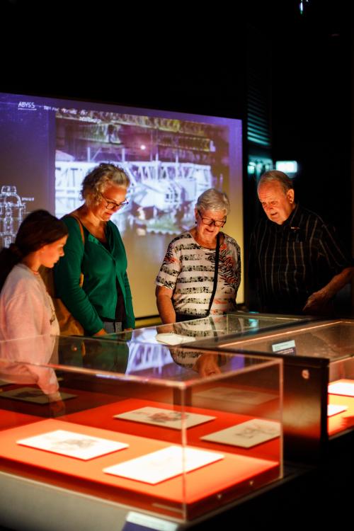 Four individuals gather around a series of glass showcases filled with paper drawings and inspect them with interest. They stand in a dramatically lit Museum exhibition space.