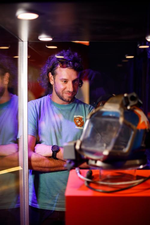 A person with short curly brown hair and a blue t-shirt stares with interest at a diving helmet on display in a dramatically-lit Museum showcase.