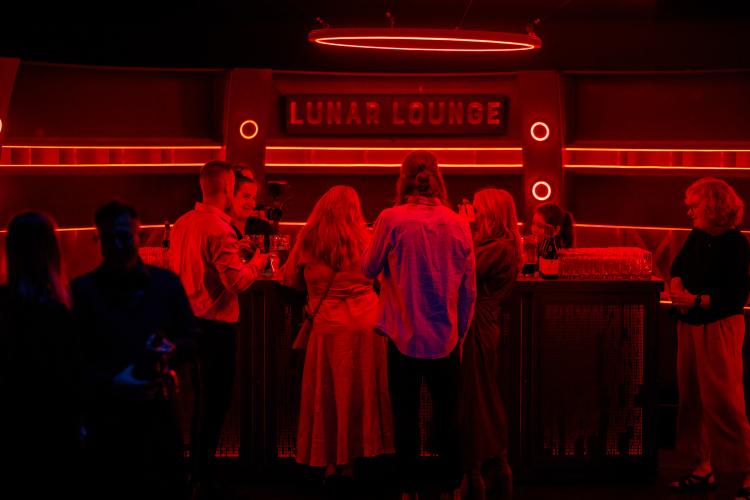 A group of people stand at a bar with their backs to the camera. The bar is decorated in a space-like sci-fi themed aesthetic and bathed in a dramatic red light.