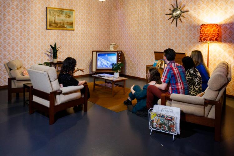 A group of people in variously coloured outfits sit on multiple cream and brown sofas and watch an old 60s style television unit. The set around them is decorated in the style in a 1960s living room style