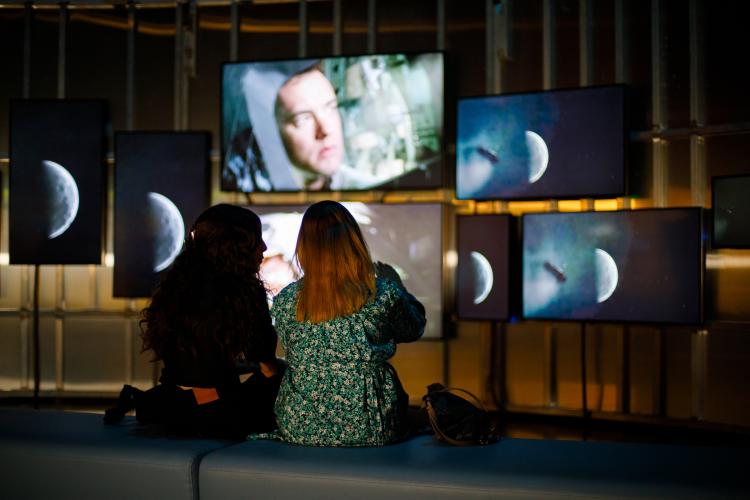 Two people sit with their backs to the camera and face a series of digital screens in various orientations showing images of the moon and an astronuat