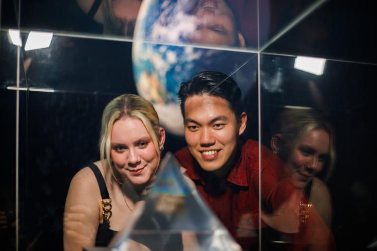 Two people stare with interest into a Museum showcase containing a glass pyramid with a moon rock inside. They stand in a dramatically-lit gallery in front of a projection of the earth.