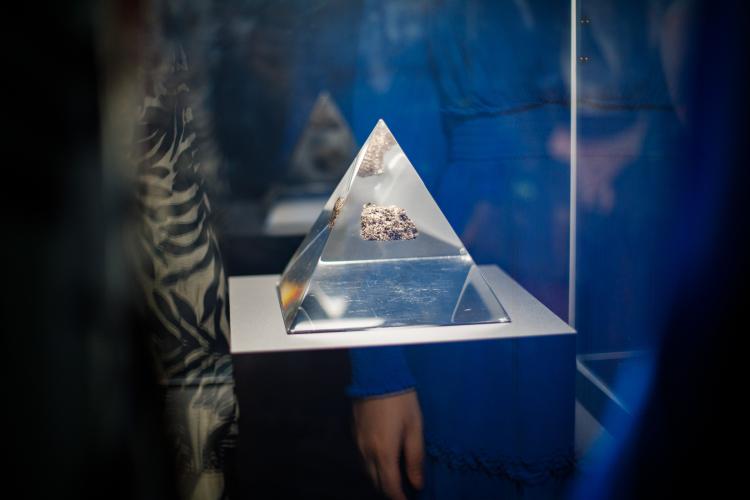 A glass pyramid structure contains a small sample of moon rock, a slightly sparkly roughly textured fragment. Reflections of onlookers are visible in the glass surface