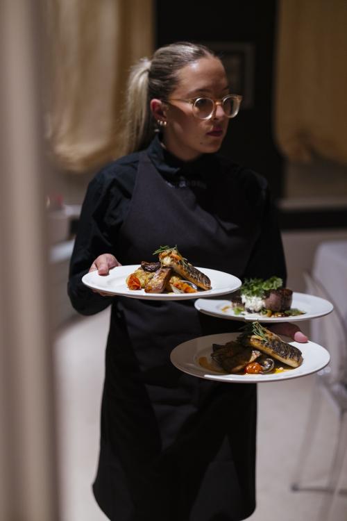 A Heyder and Shears company waitress wears a black shirt under a black apron and clear framed glasses with their hair in a pony tail. They carry three gourmet dishes of salmon and beef plated aesthetically