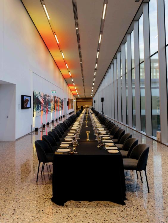 A long table and chairs set up for a function in a long hallway, surrounded by large windows and artworks