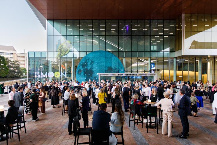 A large crowd gathers in an open area outside of the WA Museum's glass-lined entrance. People wear smart clothing and stand in groups around tables and chairs while a large graphic of a moon and astronaut is visible through the crowds