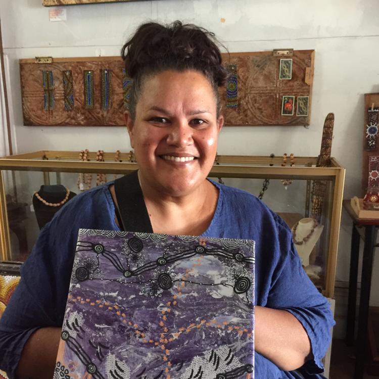 Christine Carmody wears a blue shirt with their black hair in a bun and proudly holds up a small canvas with vibrant purple and black details