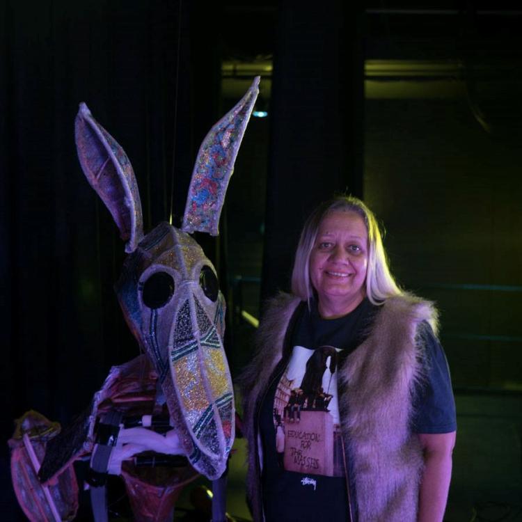Debbie Carmody wears a a black graphic shirt with a fur vest and smiles as they stand next to a sculpture of a kangaroo painted with various dot and line patterns.