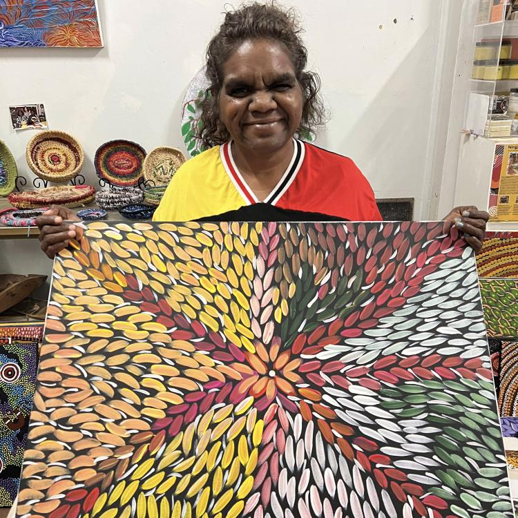 Debra Frazer wears a yellow red and black shirt and smiles proudly as they hold a large canvas painted with vivid yellow pink white and green petal-like colours.
