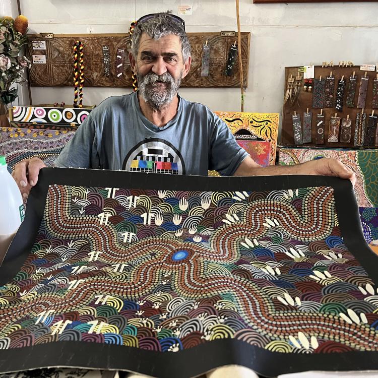 Rob Wilson has a thick black and grey beard, and wears a grey shirt. They proudly hold up a colourful painting featuring dot painted lines and concentric circles interspersed with different animal footprints