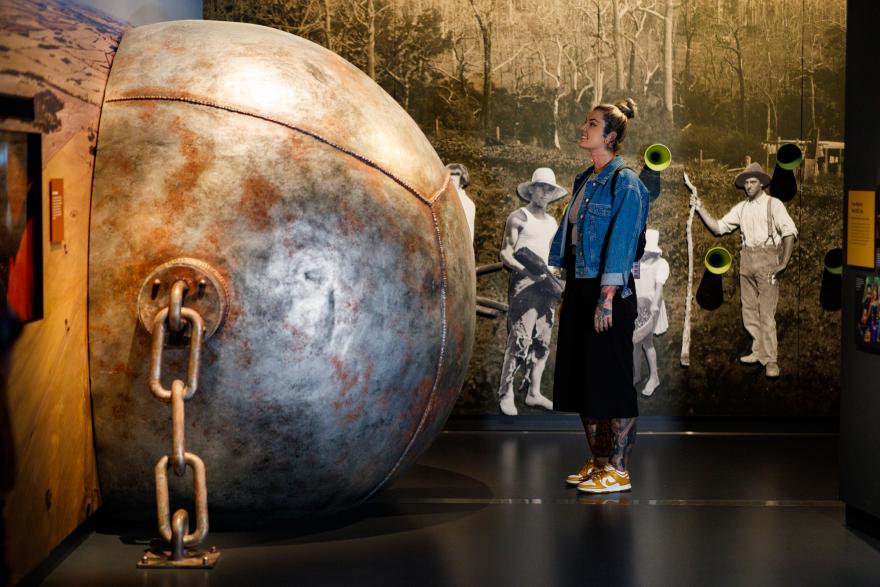 Woman in a museum looking at a large metal sphere with a chain, against a backdrop of historical worker photos.