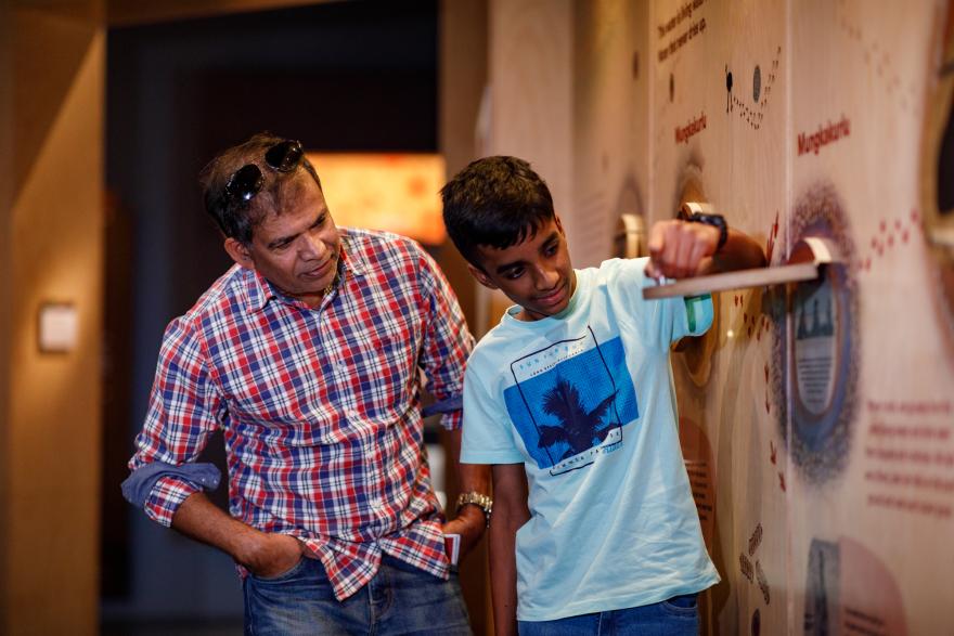 An adult male and a younger male interact with a museum exhibit, with the younger male operating a lever on the wall.