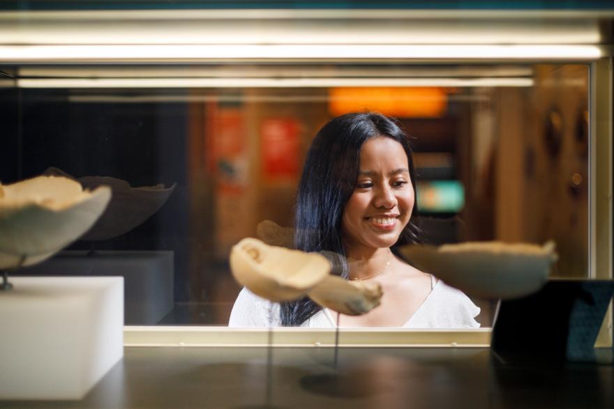 A woman smiling while looking through a museum display case containing light-colored artifacts.