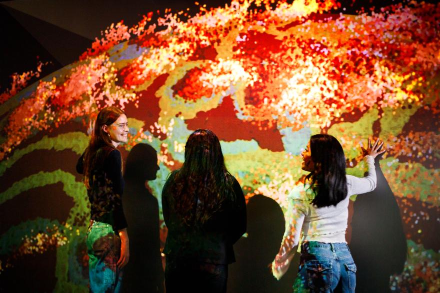 Three individuals stand in front of a vibrant, abstract, mosaic-like background with bright red, orange, yellow, green, and blue colors.