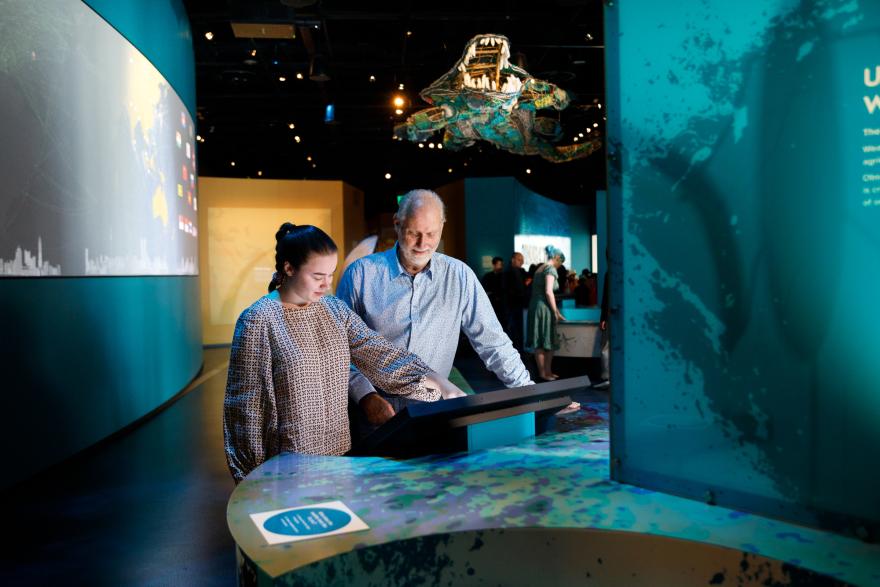 People interacting with a touchscreen exhibit in a modern, dimly-lit exhibition space with a large model creature suspended above.
