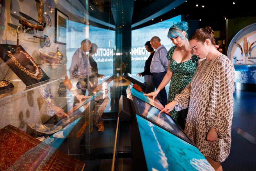 People interacting with digital displays at a museum exhibit with various artifacts displayed behind glass.