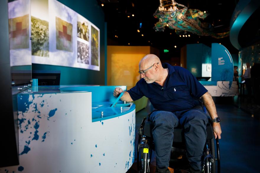 Man in a wheelchair interacting with an exhibit in a modern museum, with illuminated images and a hanging sea creature model in the background.