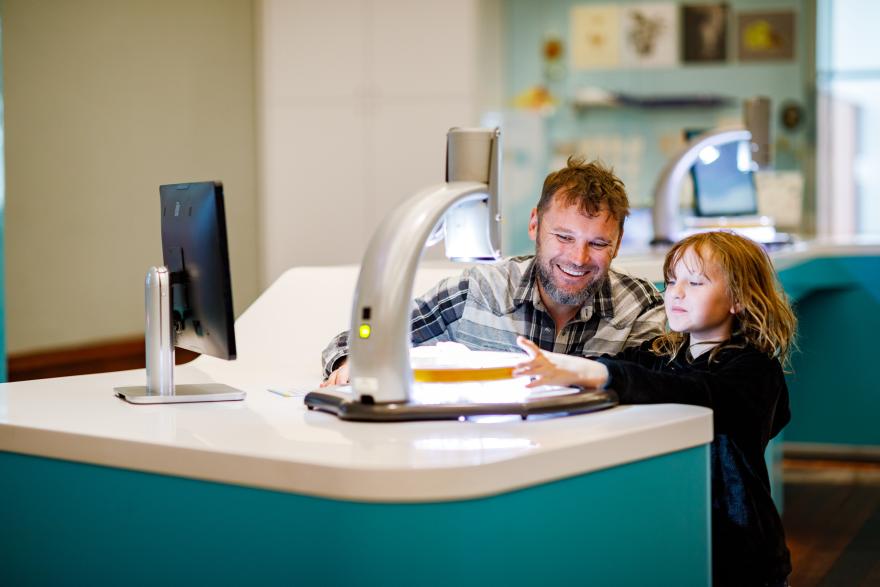A man and a young girl interact with a scanning device at a table in a modern, well-lit room.