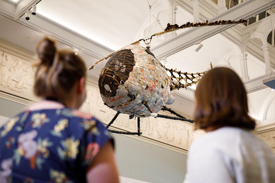 Two people from behind looking at a large sculpture made from recycled materials suspended in a museum.