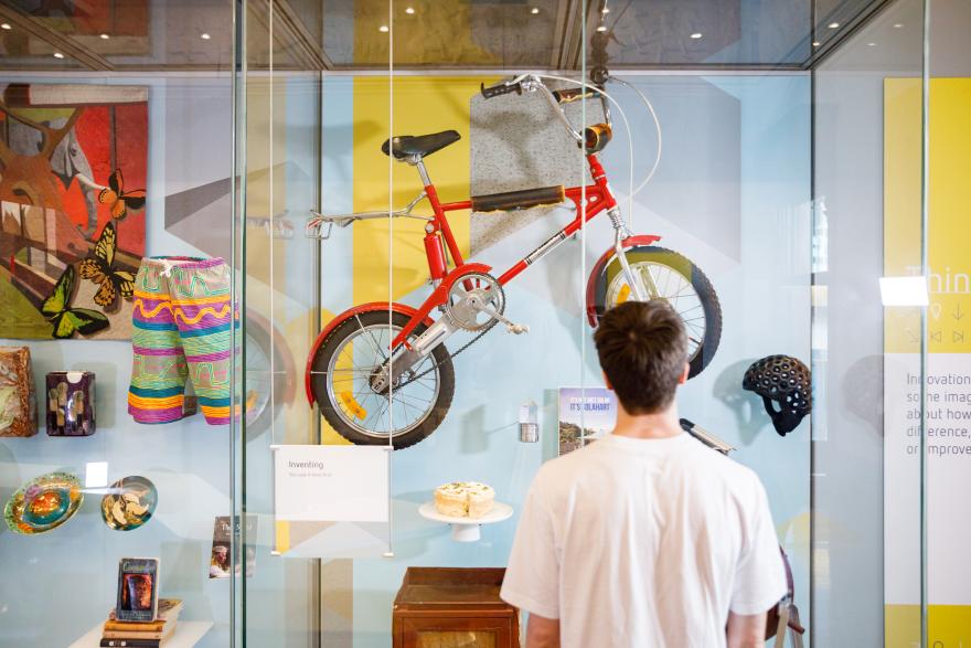 Museum display featuring a red bicycle, multicolored shorts, butterfly artwork, various decorative items, and a viewer in a white shirt.
