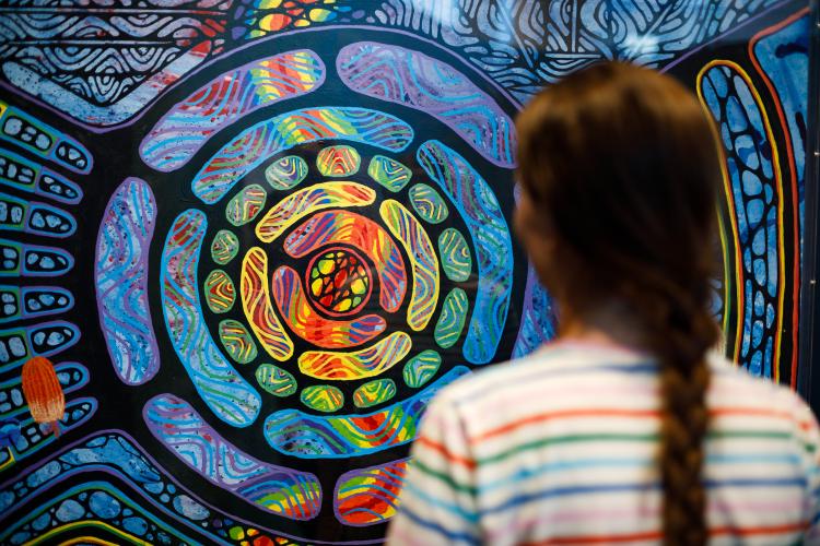 A person with a braided hairstyle observes a vibrant, abstract mural with concentric circles and colorful patterns.