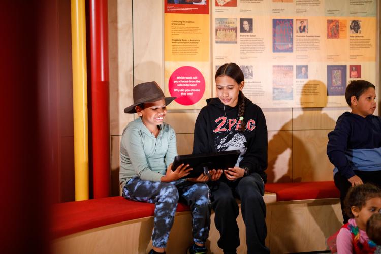 Three children sitting on a red-cushioned bench with a digital tablet in front of a wooden wall displaying book covers and descriptions.