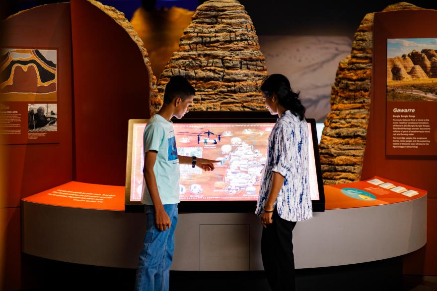 A boy and a woman interact with a touch-screen map display in a museum exhibit with rock formations and informational panels.