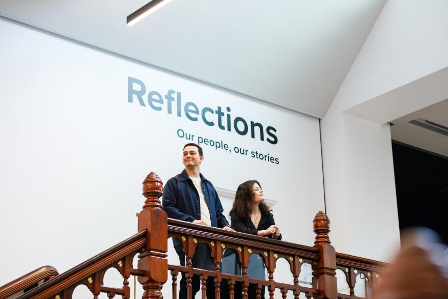 Two people stand on a wooden staircase beneath a wall with the text "Reflections: Our people, our stories."
