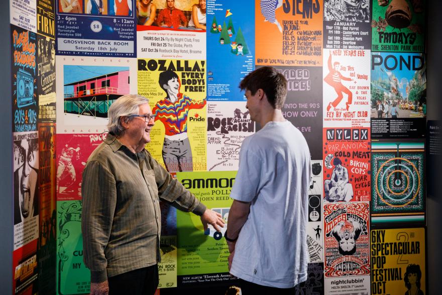 Two people conversing in front of a wall covered with colourful retro music posters.