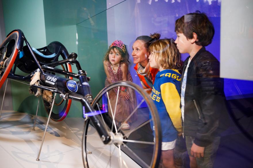 Four people, including three children, observe a racing wheelchair in a museum exhibit.