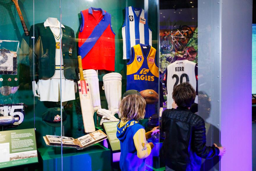 Various sports memorabilia, including cricket gear and team jerseys, displayed in a museum glass case with two children observing.