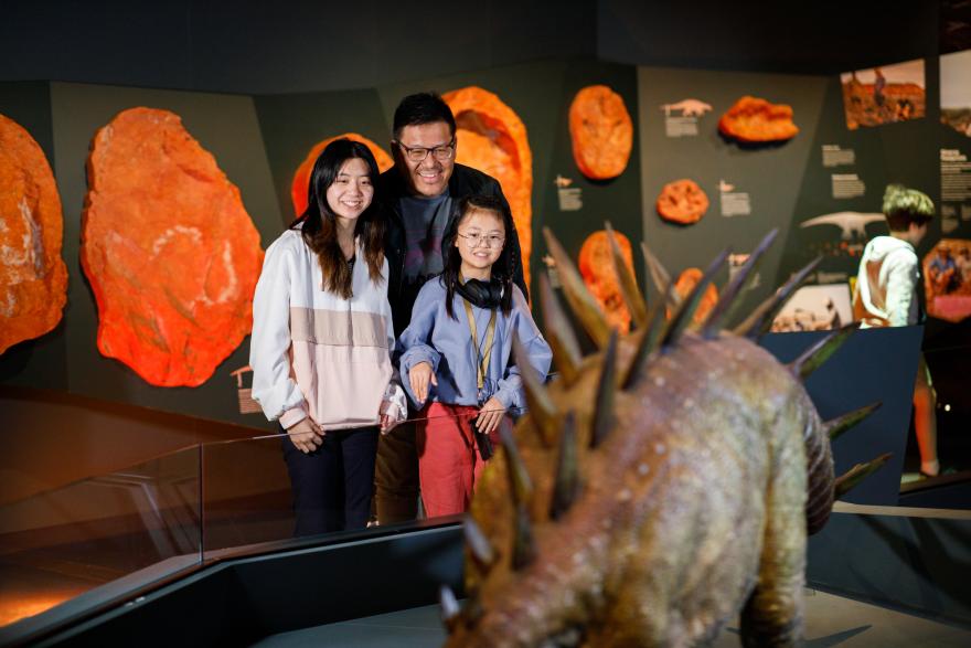Three people smiling in a museum exhibit with fossilized dinosaur tracks on the wall and a spikey dinosaur model in the foreground.