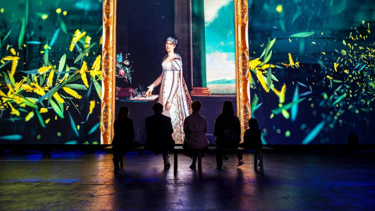 A colourful projection of a painting of Express Josephine who wears a white dress which red and blue details and a crown among images of a yellow flowering plant is projected on a long wall interspersed with the silhouettes of Museum visitors who sit on a bench watching the scene