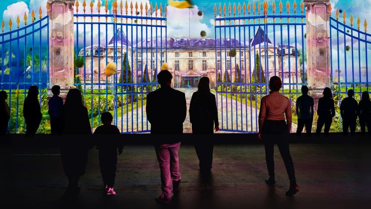 A colourful projection of an English style brick manor house and vivid green garden behind a blue and gold ornate fence on a long wall interspersed with the silhouettes of Museum visitors who stand watching the scene