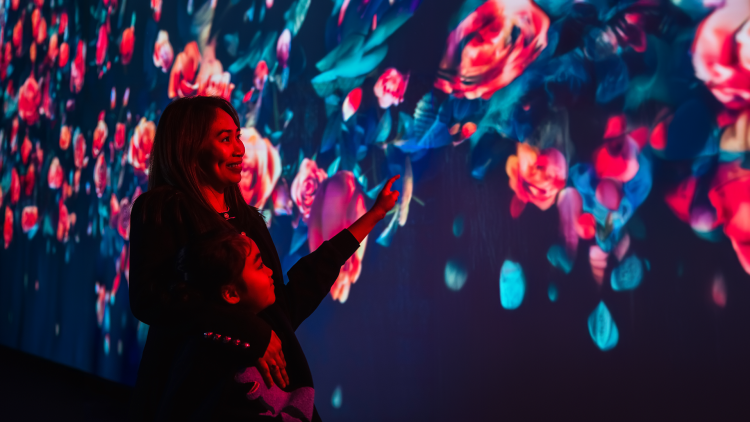 An adult and a young child stand in front of a large colourful projection of roses and leaves. The young person reaches out their hand to point at the projection