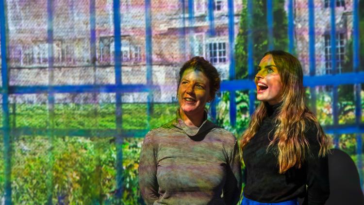 Two people, one in a striped shirt and tied up brown hair, and the other in a black shirt with their long brown hair worn down stare with expressions of excitement at something off-camera. A vividly colourful projection of an English garden and manor house behind a blue fence is projected over the people and onto the wall behind them.