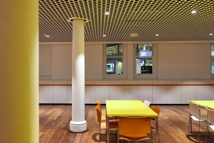 A modern working area with yellow tables, orange and white chairs, wooden floors, white columns, honeycomb-patterned ceiling, and windows in the background.