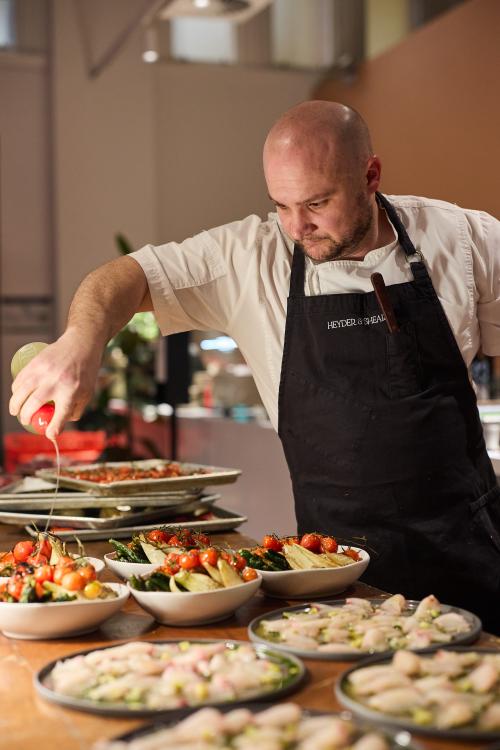 Chef in a professional kitchen pouring a dressing over several plates of vibrant salads.