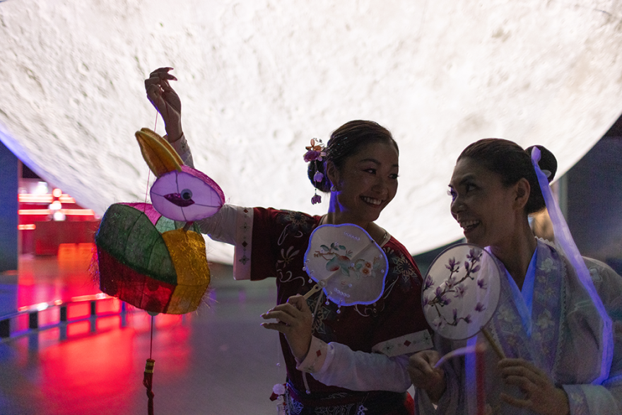 2 people looking at each other with a large display moon behind them, holding a rabbit lantern and traditional fans