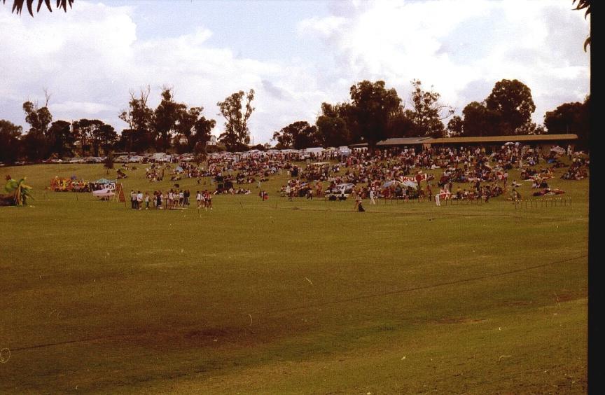 Large outdoor field with crowds of people, trees in the background, and cloudy sky overhead at a public event.