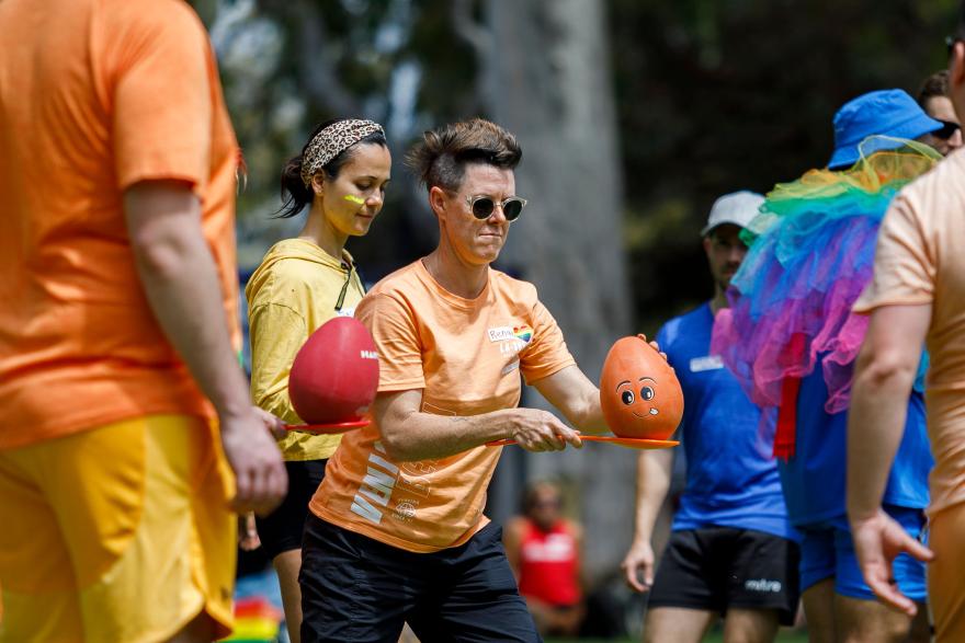 A group of people outdoors, one balancing an egg on a spoon, surrounded by colourful clothing and a tree background.