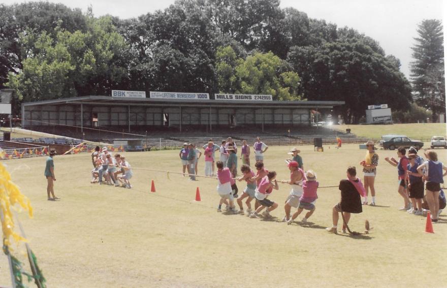 Outdoor tug-of-war event with teams pulling a rope on a grassy field, surrounded by spectators and trees.