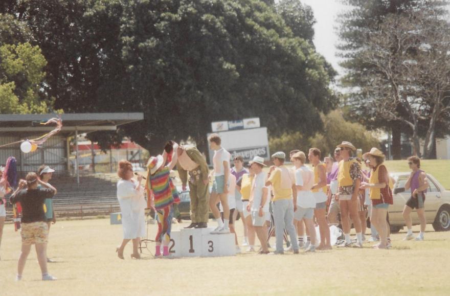 Outdoor event with people on a podium and a line of participants waiting.