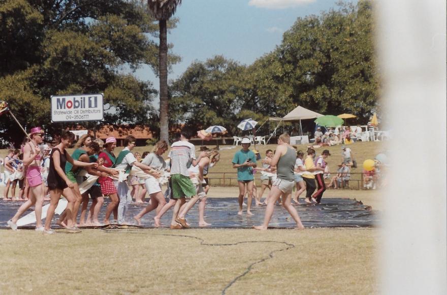 People participating in an outdoor water sports event on a grassy field with a large tree and umbrellas in the background.