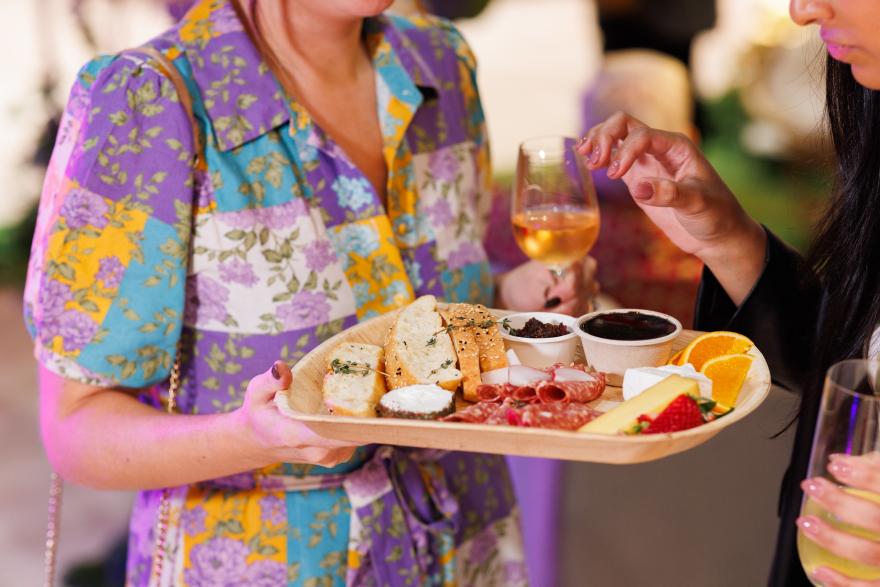 Two people holding a wooden platter with various foods, including bread, cheese, fruit, and two glasses of wine.